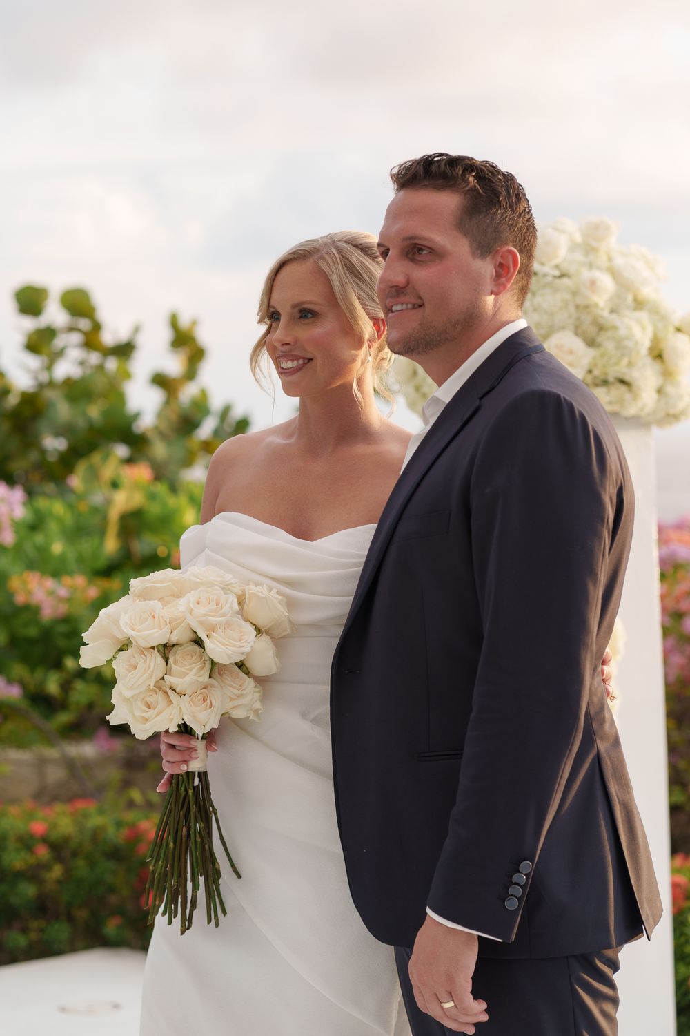 A romantic outdoor wedding scene with the bride in a white strapless gown holding white roses and groom in dark suit.