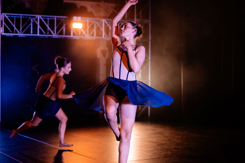 Dancers perform a dynamic duet under stage lighting with one performer in a blue tutu making a graceful leap.