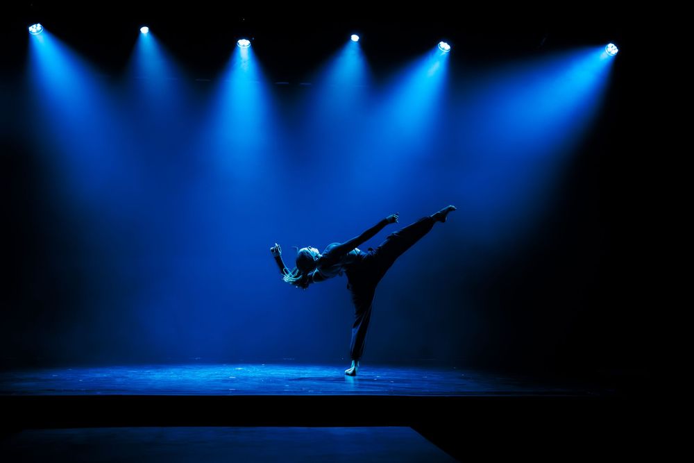 Modern dancer silhouetted against dramatic blue spotlights performs an arabesque on a dark stage.