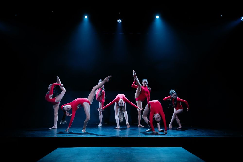 Group of dancers in red costumes perform a contemporary piece under blue stage lighting.