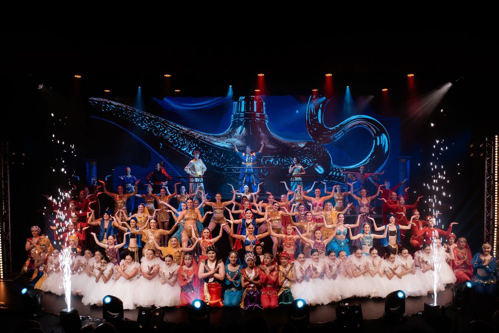 Stage performers in colorful costumes pose together during grand finale with pyrotechnics and dramatic blue lighting effects.