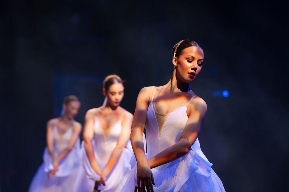 Ballet dancers perform elegant choreography in flowing white dresses against a dark background.