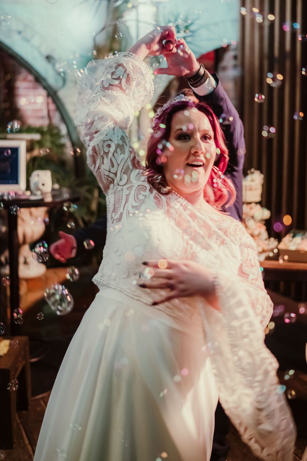A person in a flowing white wedding dress joyfully plays with soap bubbles during a celebratory moment at an indoor venue.
