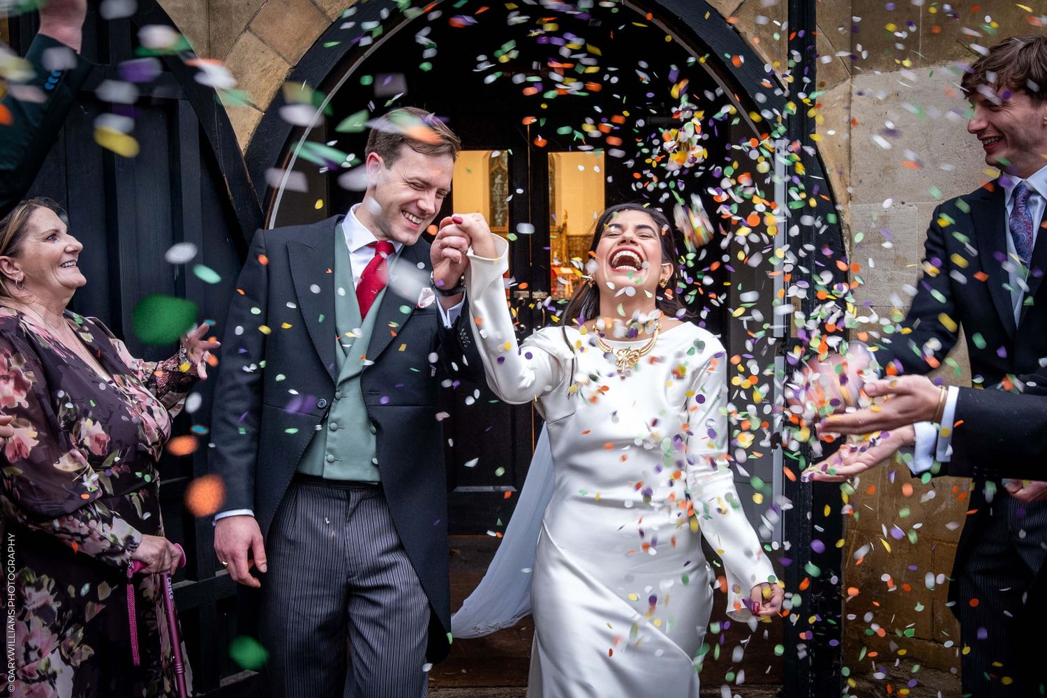 Joyful newlyweds celebrate under shower of confetti at church doorway.