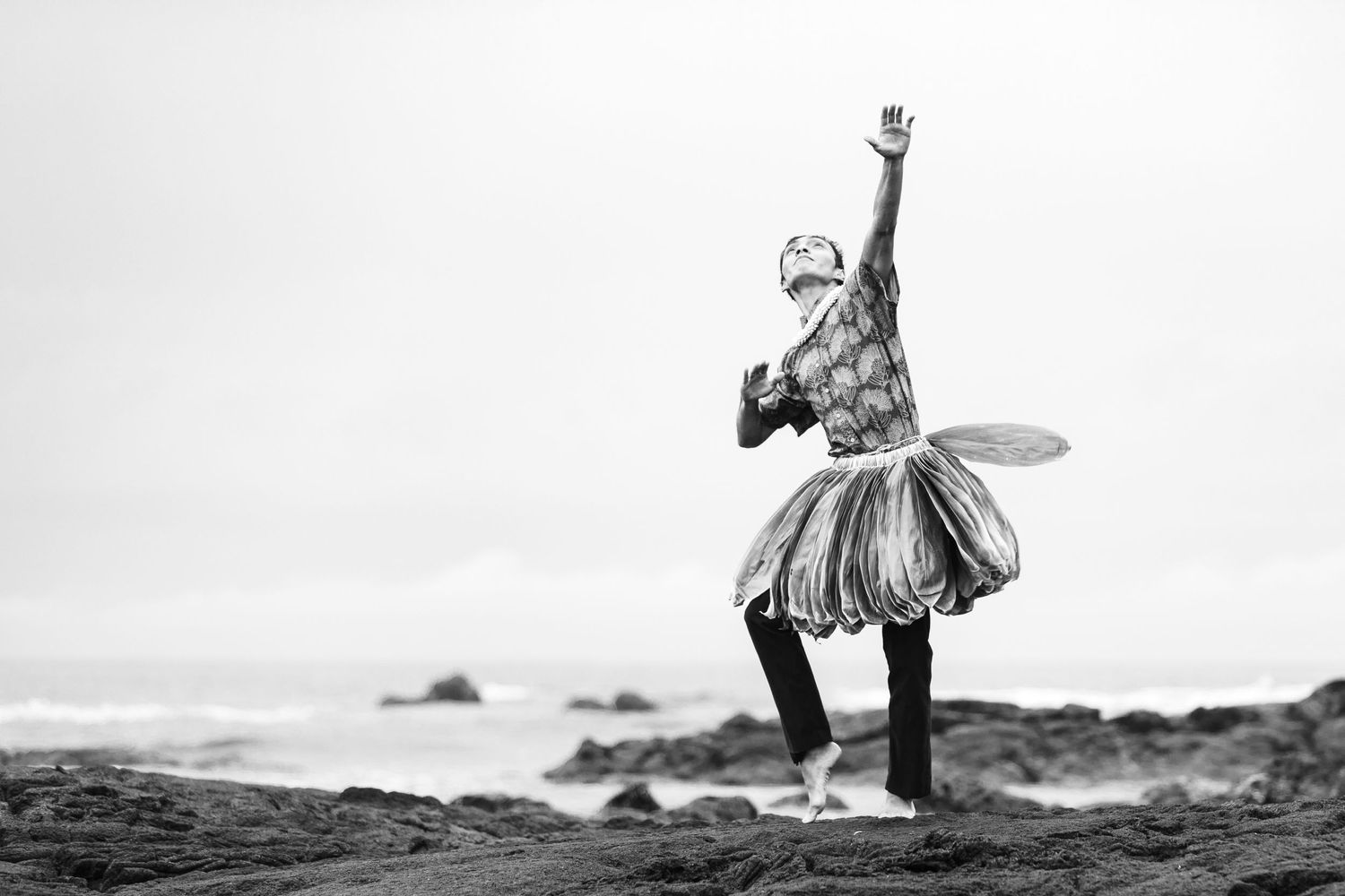 A dancer in a tutu performs an elegant ballet pose against a dramatic rocky coastline in black and white.