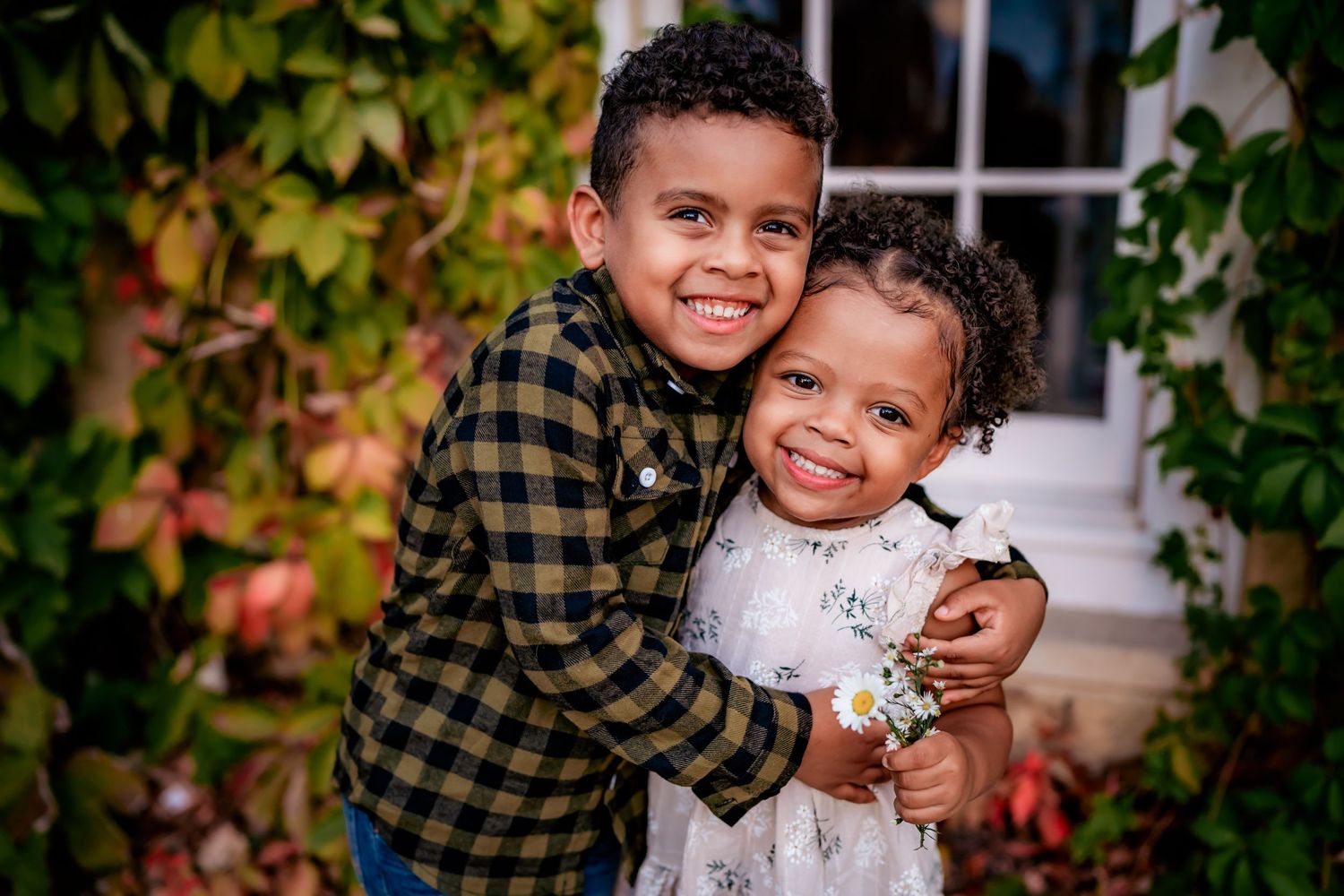 Two children embrace and smile warmly while standing together in front of an autumn-decorated porch with climbing vines.