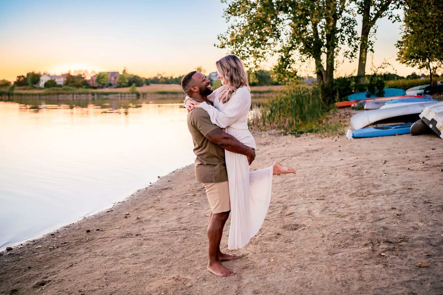 A couple embraces on a sandy beach at sunset with boats in the background near a tranquil lake.