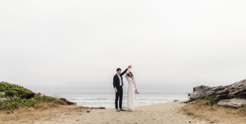A couple in wedding attire stands together on a misty coastal cliff overlooking the ocean.