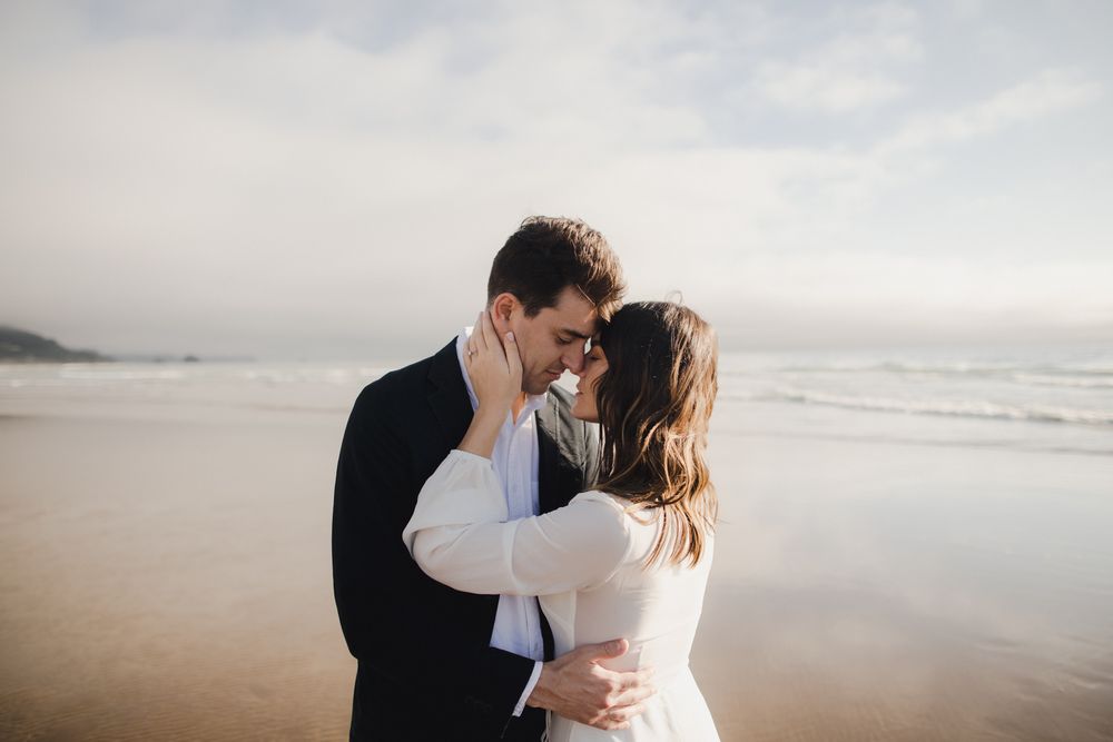 A romantic couple shares an intimate moment on a misty beach at sunset, dressed in elegant black and white attire.