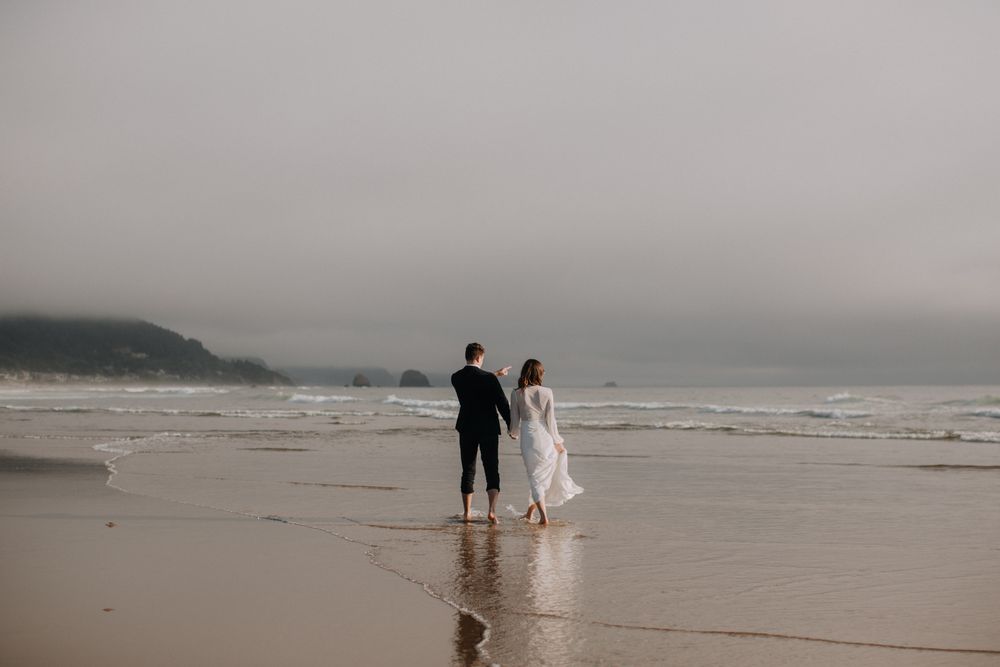 A couple walks hand in hand on a misty beach with dramatic overcast skies and crashing waves in the background.
