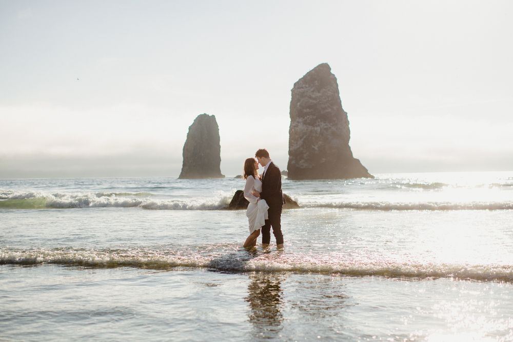 Silhouetted couple stands in ocean waves with dramatic rock formations at Cannon Beach, Oregon at sunset.