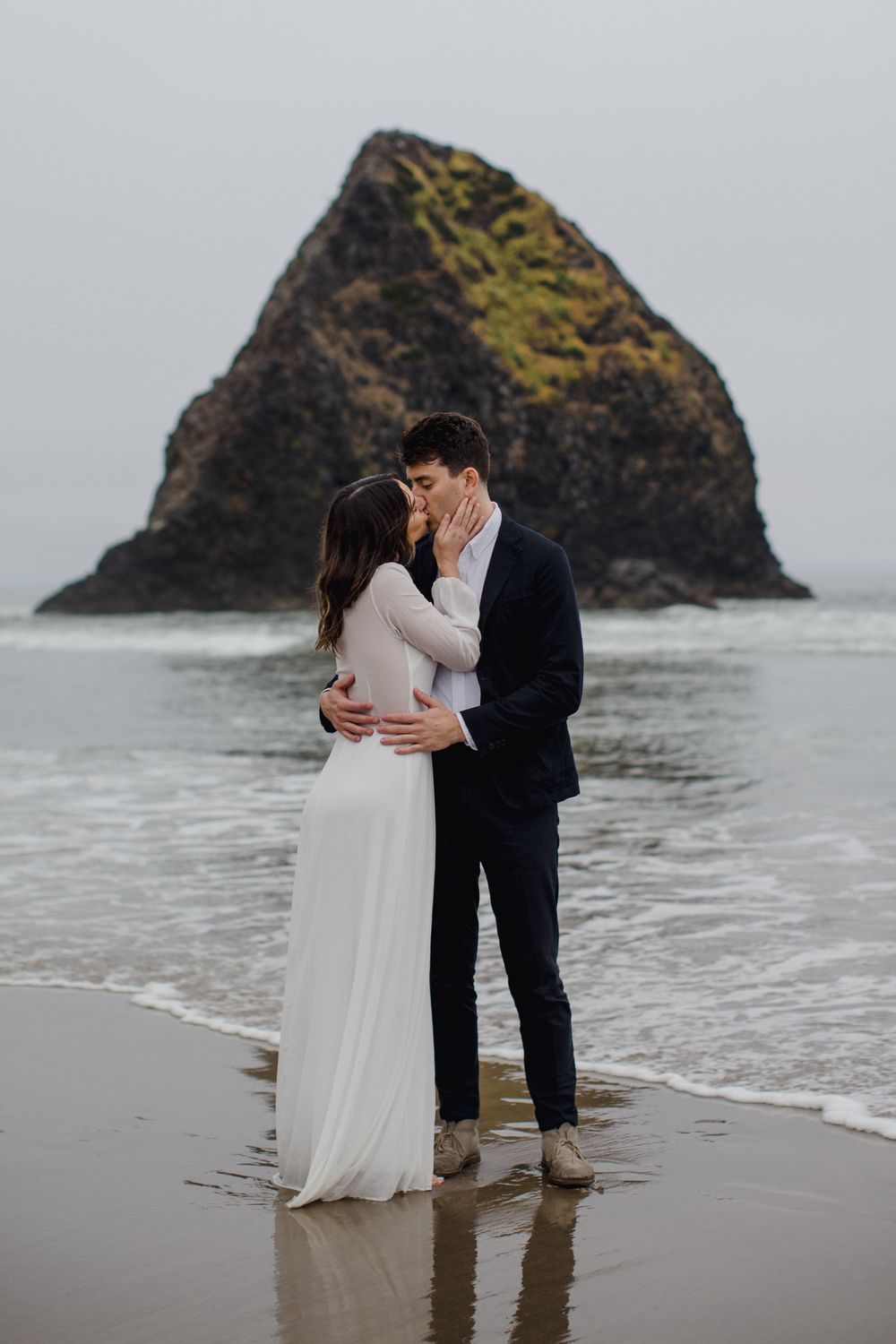 A romantic couple shares an intimate moment on a misty beach with iconic Haystack Rock at Cannon Beach in Oregon.