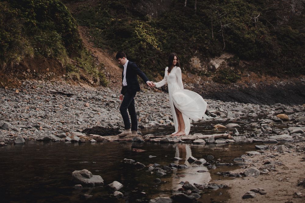 Two wedding attired figures holding hands while standing on rocky beach shore with reflections in water at dusk.