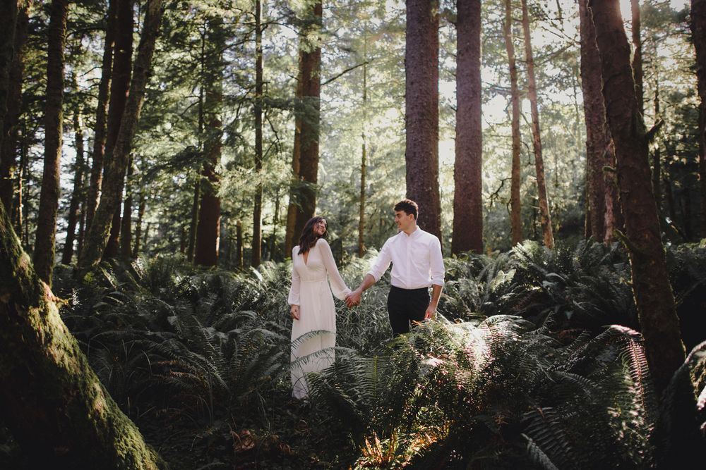 A couple standing together in a sunlit forest with tall trees and lush ferns surrounding them.