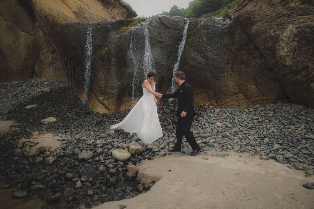 A couple in wedding attire dance together on a rocky beach beneath a dramatic waterfall cascading down cliffs.
