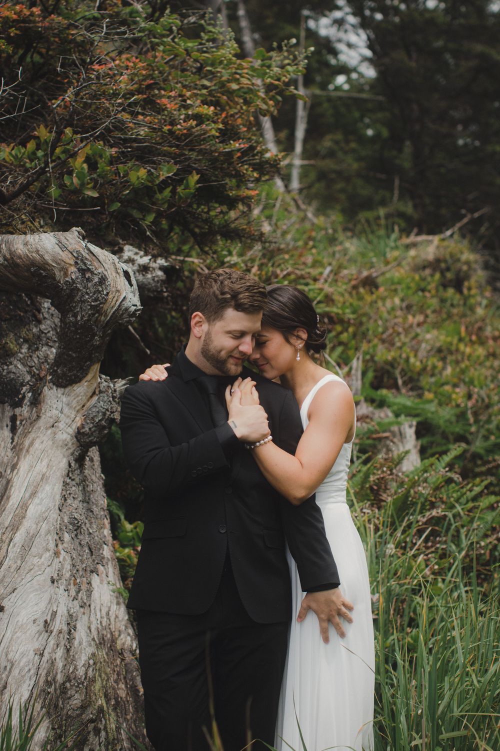 A romantic wedding couple embraces near a fallen tree trunk in a lush forest setting during autumn.
