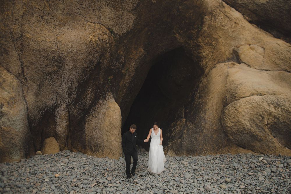 Two people in wedding attire walk together on a rocky beach near dramatic cave formations at sunset.