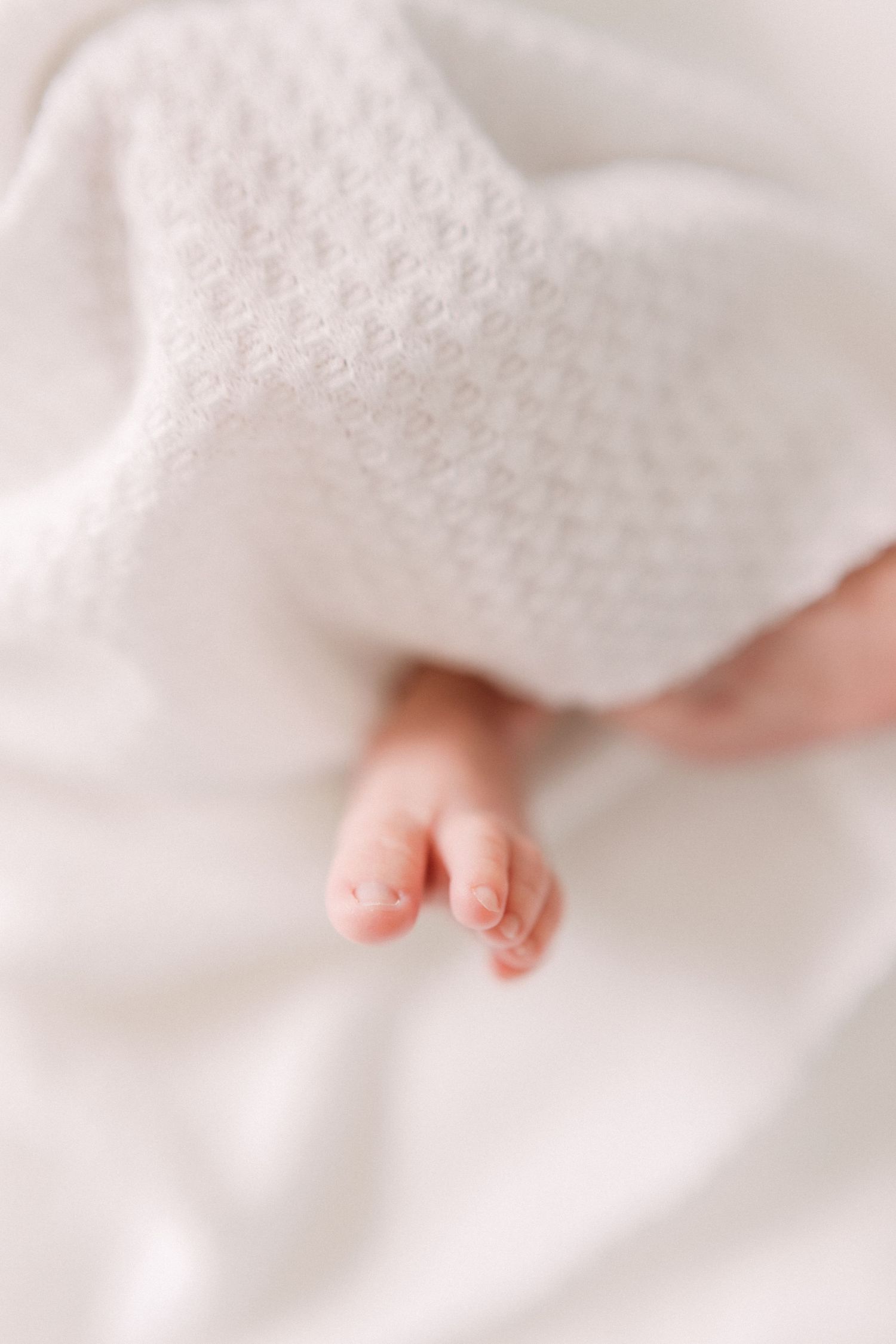 Tiny baby foot peeking out from soft white blanket in dreamy, gentle lighting.