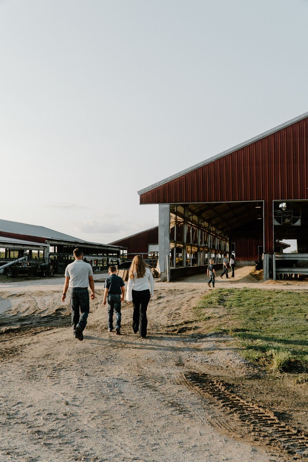 Three people walk towards a large red barn building on a dirt path at a dairy farm.