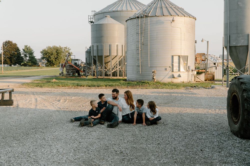 A group sits together on gravel near large grain silos at a dairy farm during golden hour.