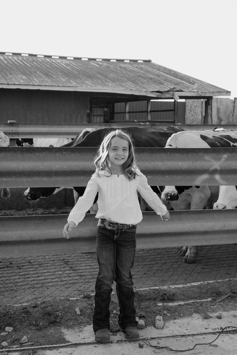 A young person stands in front of farm fencing with cattle visible in the background.