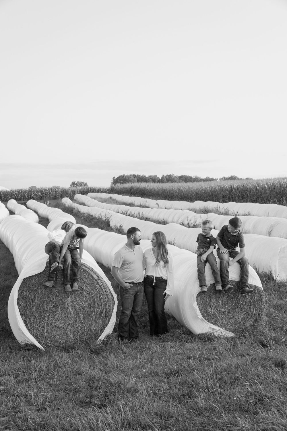 Family members pose on large wrapped hay bales in a rural field setting.