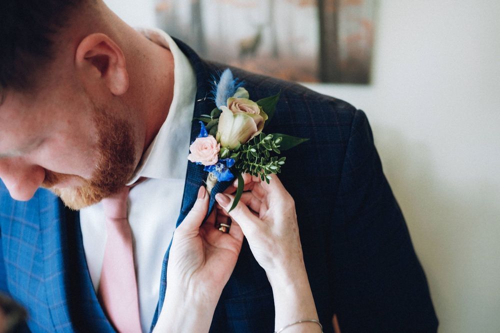 A groom getting ready on his wedding day adjusts his boutonniere while wearing a navy suit and light blue tie.
