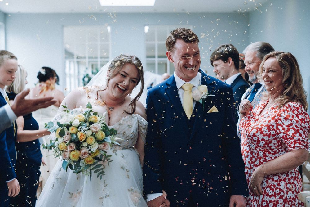 Wedding guests shower newlyweds with confetti as the bride holds a yellow and white bouquet outside.