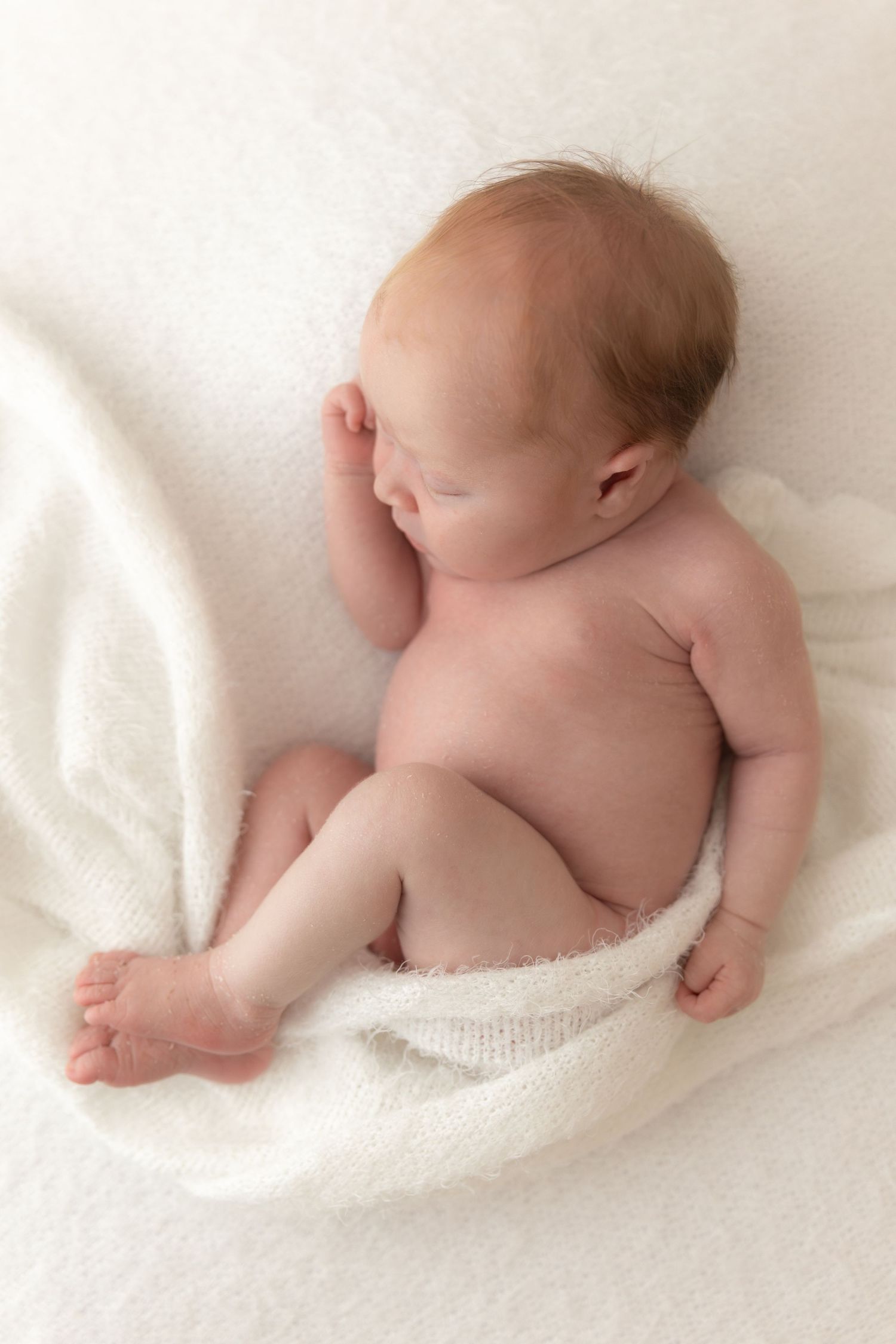 A sleeping newborn baby lies on a soft white blanket with bare feet and torso exposed.
