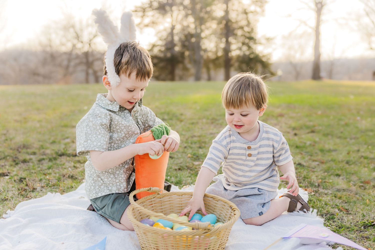 Two children playing with Easter eggs on a white blanket in a sunlit park.