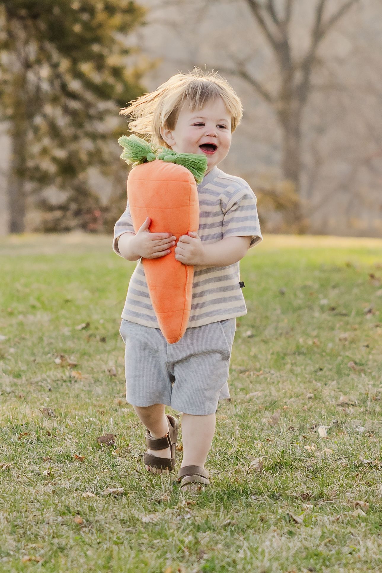 Young child running with a plush carrot toy while laughing in a grassy field. Mini session.
