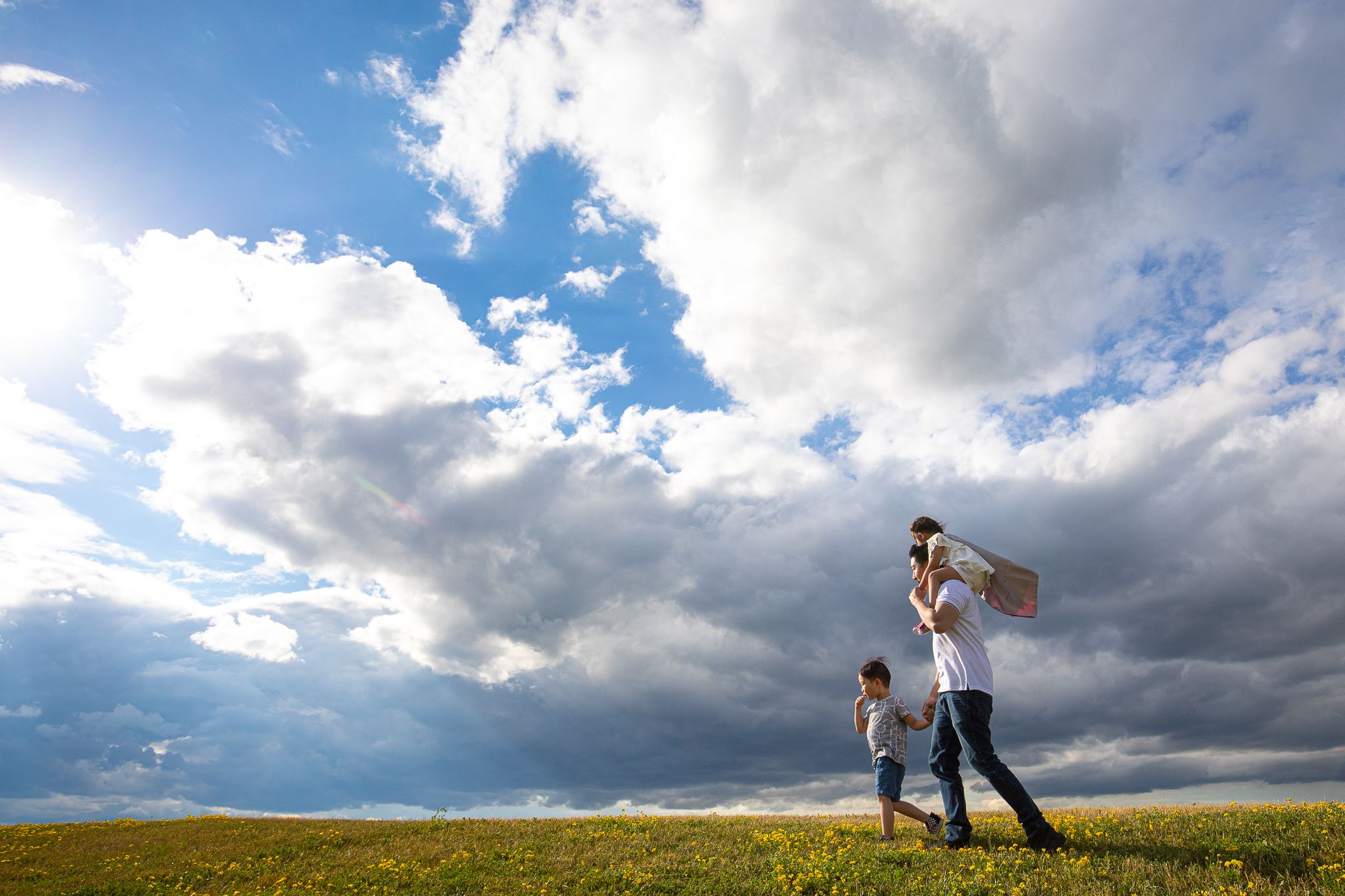 Two people walk across a green field under dramatic cloudy skies with sunbeams breaking through on a summer day.