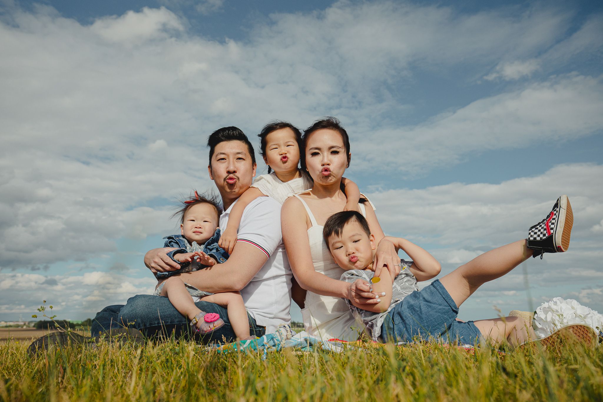 Playful family poses together on grassy field against cloudy blue sky backdrop.