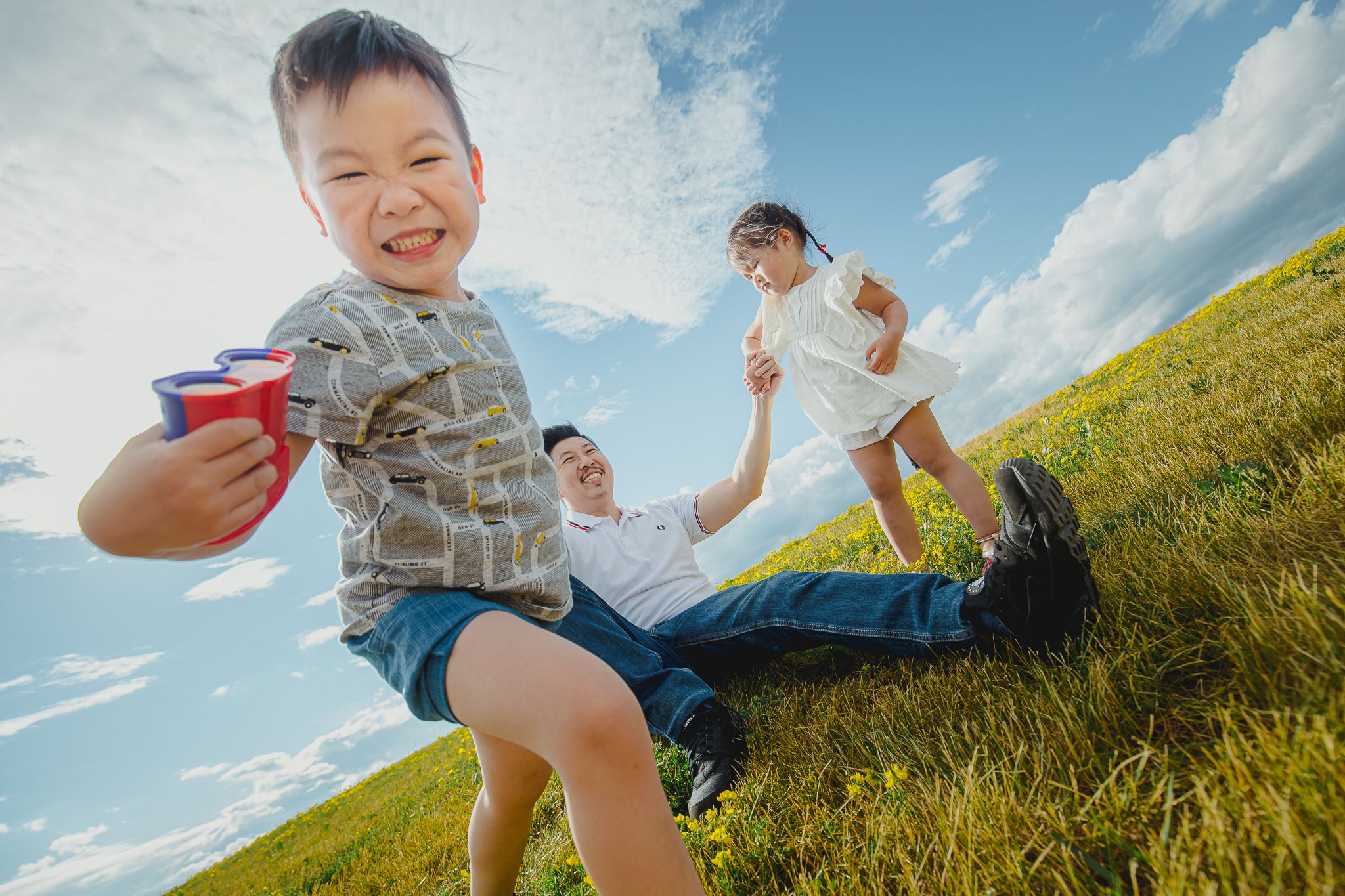 A family enjoying a playful moment on a grassy hillside under bright blue summer sky.