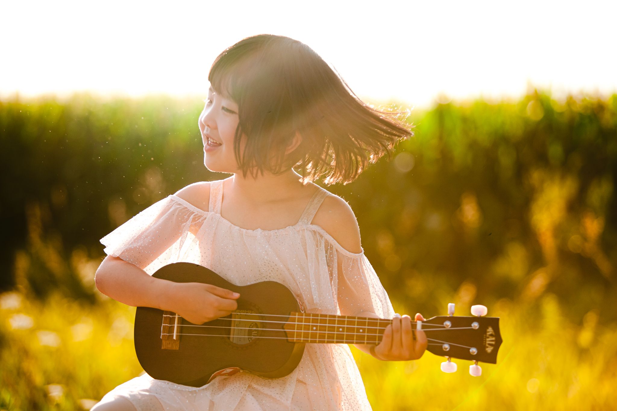 A person in a white dress plays ukulele while sitting in a sunlit meadow during golden hour.