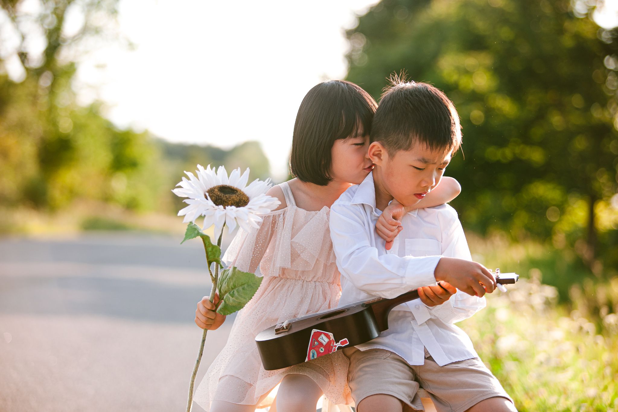 Two children sitting on a road while holding a white daisy flower on a sunny summer afternoon.