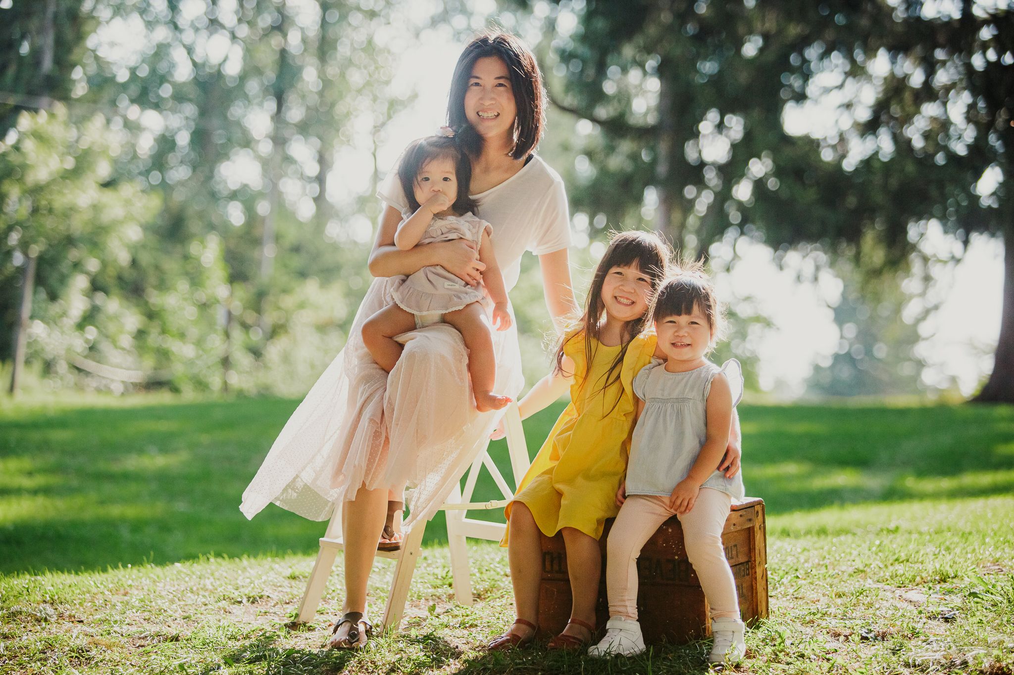 A family portrait outdoors on a sunny day with children sitting on vintage suitcases in a grassy park setting.
