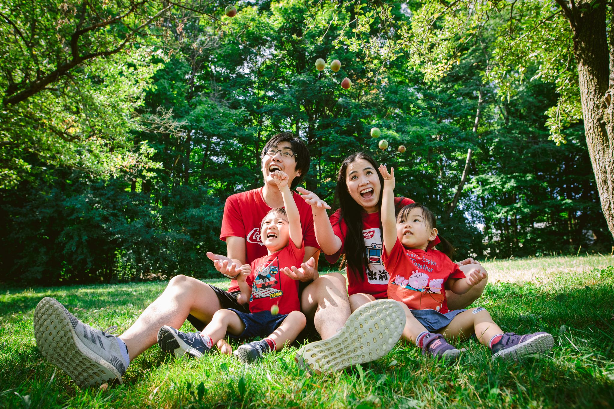 Family wearing red shirts enjoys a summer picnic together on grassy lawn beneath shady trees.