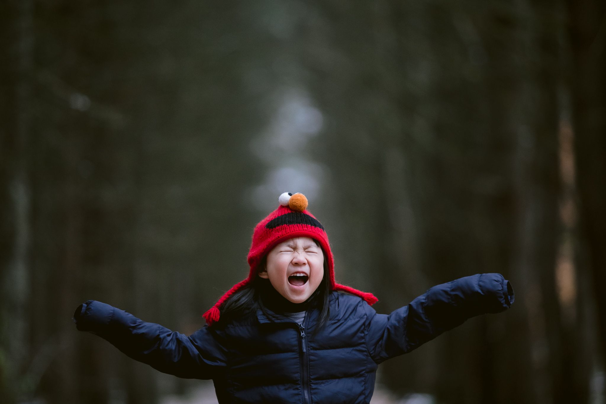 A child in winter clothing spreads their arms wide with joy in a dark forest setting, wearing a red hat and black coat.