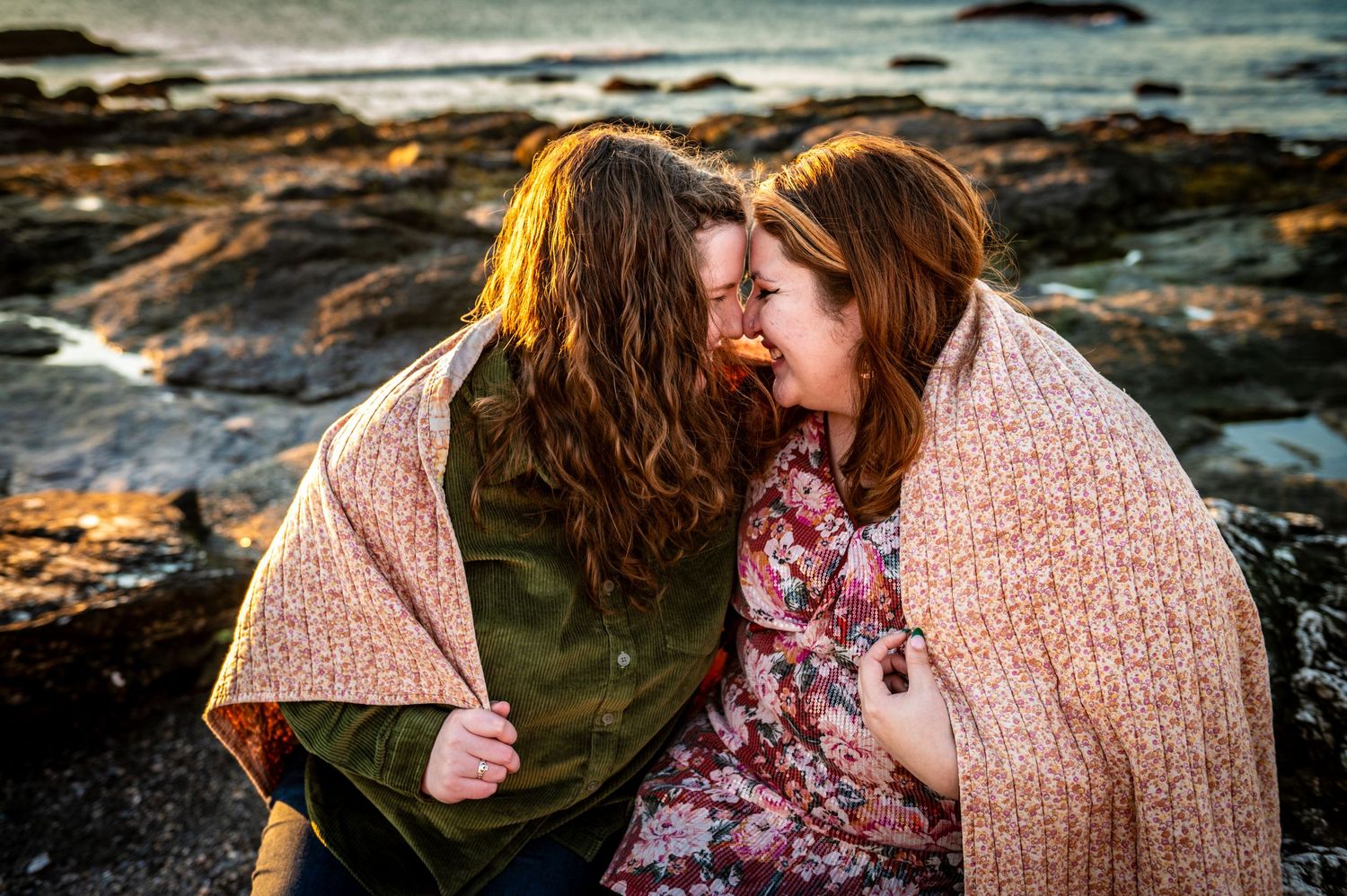 Two people wrapped in blankets share an intimate moment on rocky coastal shore at sunset.