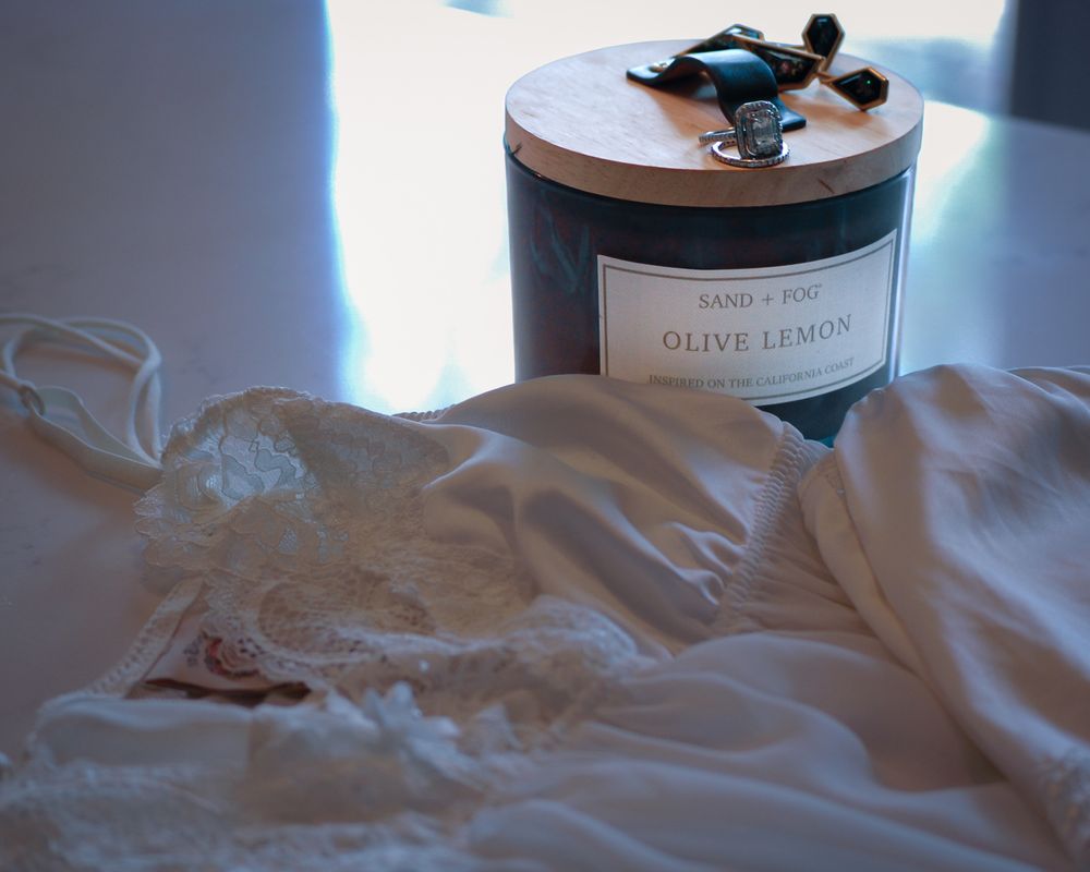 White candle jar labeled Olive Lemon placed on soft white fabric with decorative elements. Product photography, San Diego, California
