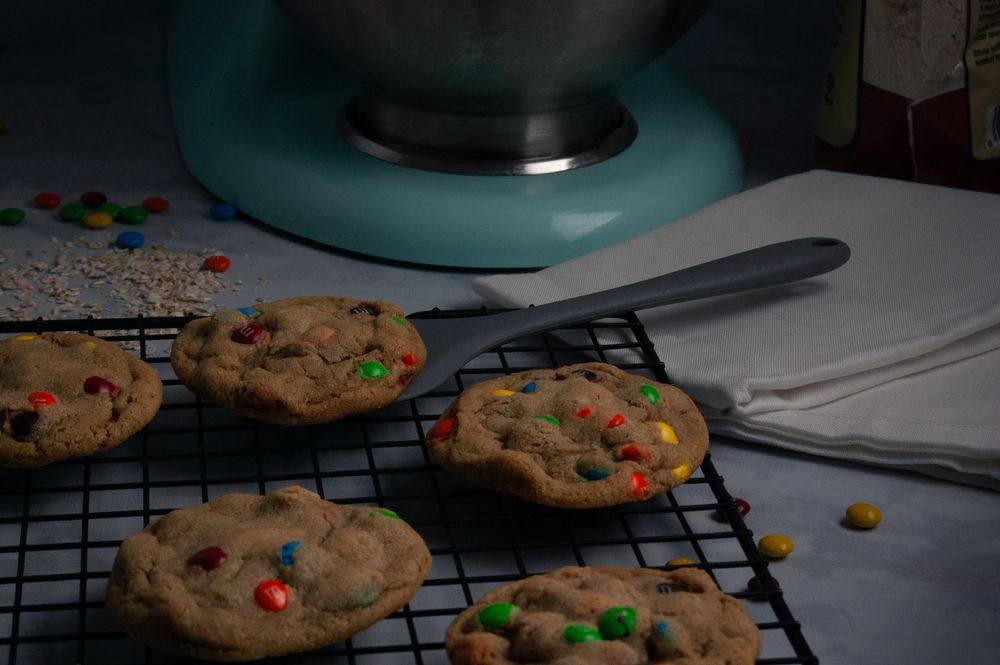 Freshly baked cookies with colorful M&Ms cooling on a wire rack near a turquoise mixer. Product photography, San Diego, California