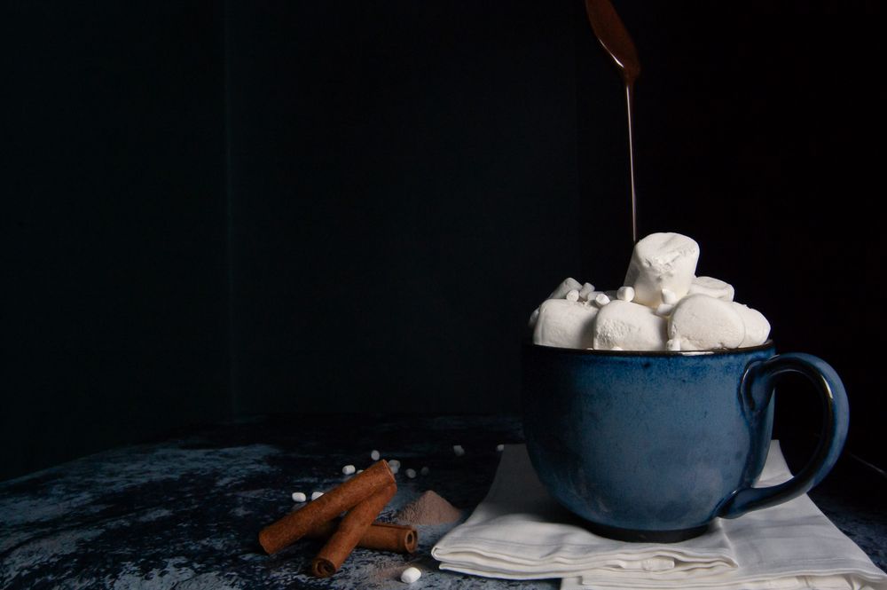 Hot chocolate pouring over marshmallows in a blue mug with cinnamon sticks. Product photography, San Diego, California
