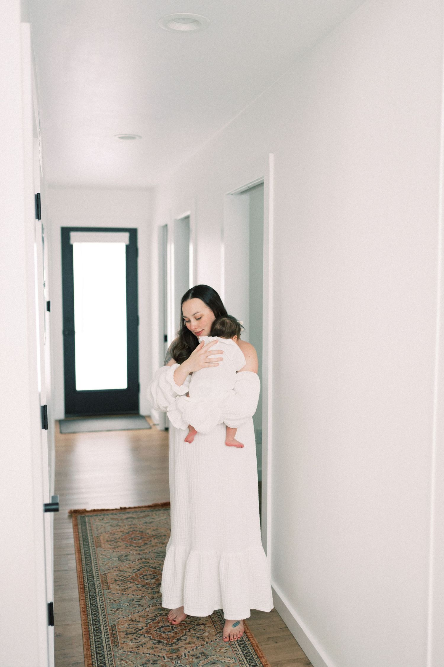 A mother in a long white dress holds her baby while standing in a minimalist hallway with a vintage rug.
