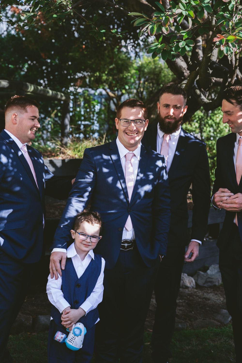 Wedding party stands together in navy suits under dappled sunlight.