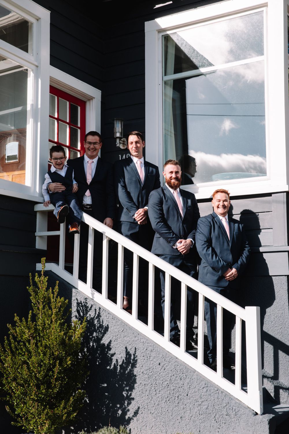 Groomsmen in suits posing on white porch stairs of a black house with red door.