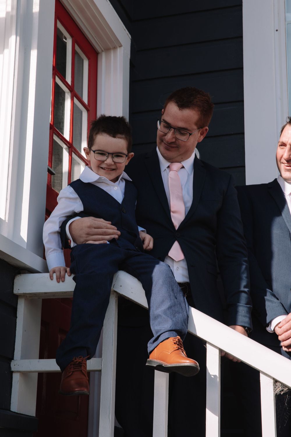 A group of men in formal attire smiling together on a white porch with black siding.