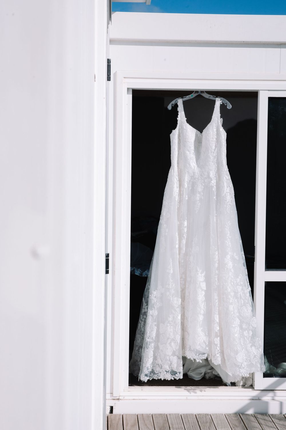 White lace wedding dress hanging in window against light background.