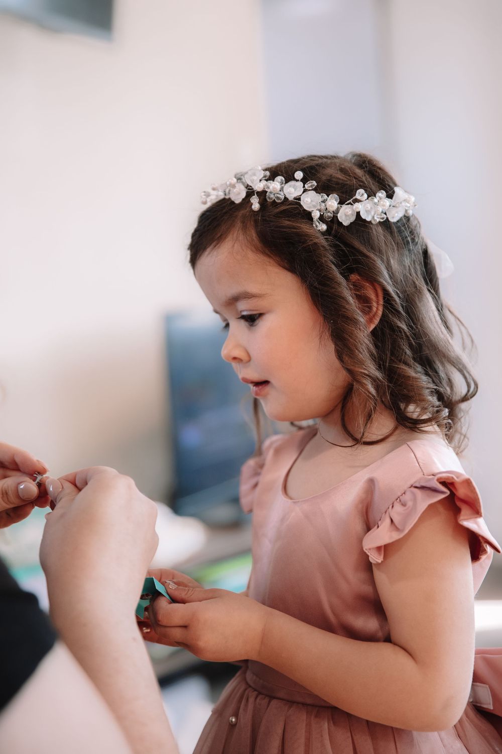 A young flower girl in a pink dress and floral headpiece looking down at something being shown to her.