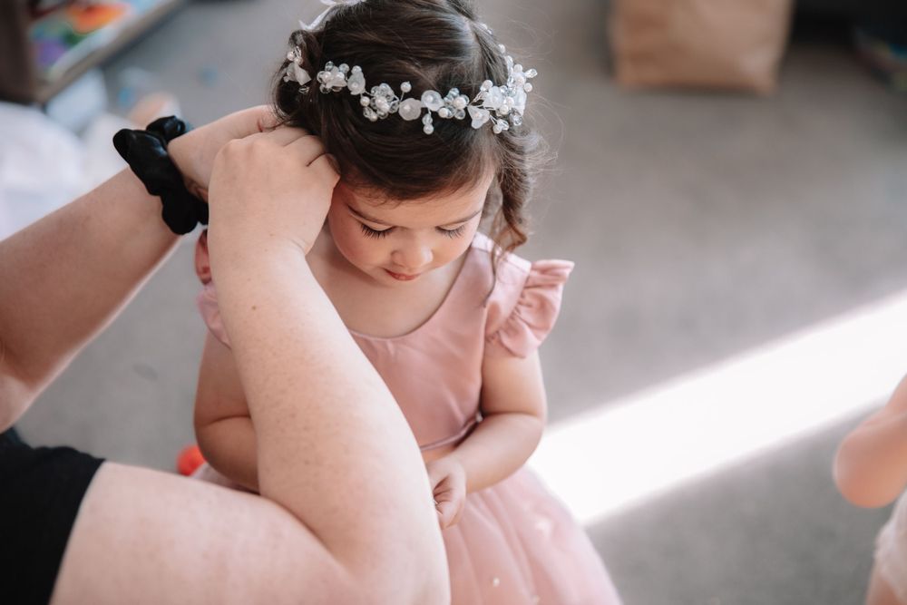 A child in a pink dress and floral crown looking down while someone adjusts their hair.